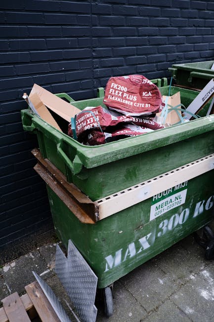 A worker wearing a yellow and red high-visibility uniform is performing rubbish removal tasks at the rear of a large, red municipal waste collection vehicle parked on the roadside. The vehicle's back 