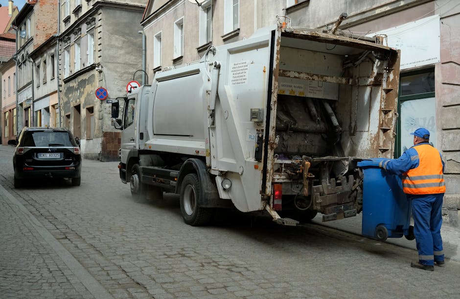 A small, old blue pickup truck with a black cargo bed is parked on a street, fully loaded with a mix of discarded cardboard boxes, rubbish bags, and miscellaneous waste materials spilling over the sid